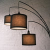 Trilogy 3-Light Floor Lamp featuring three staggered drum shades. The black woven fabric texture is clearly visible. Elegant curved arms extend from a sturdy central pole. Functional ambient lighting for reading or relaxing.