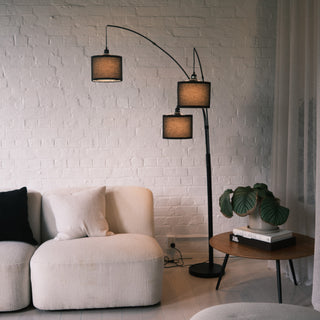 Trilogy 3-Light Floor Lamp standing in a modern living room setting. Features three arc arms and textured fabric shades. Illuminates a cream sofa and wooden side table. The white brick background enhances the matte black finish.