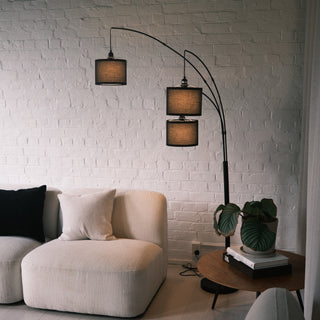 Close view of the Trilogy 3-Light Floor Lamp's staggered drum shades. The black woven fabric creates a soft, ambient glow against a white brick wall. Highlights the elegant arched metal construction and distinct black borders on the shades.