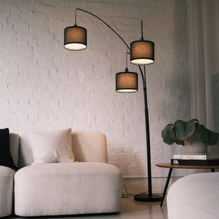 Trilogy 3-Light Floor Lamp framed by natural green foliage. Features three drum shades in textured black fabric. The curved multi-arm design adds vertical dimension to the room. Placed in a nature-inspired interior setting.