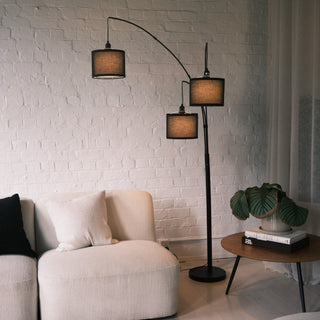 Trilogy arc floor lamp with a sturdy black metal base and three sweeping stems. Features black fabric shades with a textured weave. Positioned to illuminate a modern cream sofa and wooden coffee table. Sleek design suitable for contemporary living spaces.
