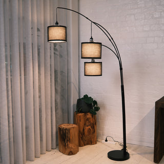 Trilogy 3-light arc floor lamp featuring a matte black metal finish and curved arms. Three textured fabric drum shades are suspended at staggered heights. Shown beside rustic wooden log tables and sheer curtains. The woven shades diffuse light for a soft, warm atmosphere.
