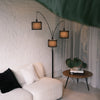 Trilogy 3-light arc floor lamp featuring matte black metal stems. Three textured fabric drum shades with distinct black borders hang at staggered heights. The woven material diffuses light for a warm, ambient glow. Durable multi-arm design. Positioned in a modern living space with a white brick wall.