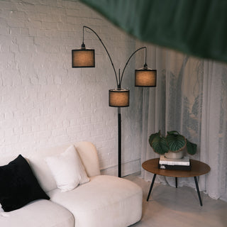 Trilogy 3-light arc floor lamp featuring matte black metal stems. Three textured fabric drum shades with distinct black borders hang at staggered heights. The woven material diffuses light for a warm, ambient glow. Durable multi-arm design. Positioned in a modern living space with a white brick wall.