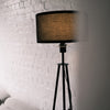 Close view of the Vanta floor lamp with its black drum shade softly lit, positioned beside a white brick wall and partially framed by a blurred curtain for a warm, modern look.