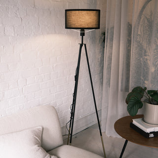 The Vanta floor lamp featuring a black KD fabric drum shade and matte black metal legs with gold dipped ends, placed beside a cream sofa and a wooden side table with a potted plant. Soft curtains and a white brick wall create a warm, modern living space illuminated by the lamp’s ambient glow.