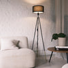 A sleek black Vanta floor lamp with a drum style KD fabric shade, matte black metal frame, and gold dipped leg ends. The lamp stands beside a soft cream lounge chair and a wooden side table with books and a potted plant, styled against a white brick wall to show its modern and warm ambient lighting effect.