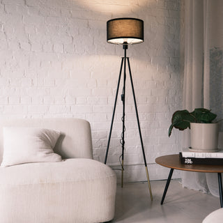 A sleek black Vanta floor lamp with a drum style KD fabric shade, matte black metal frame, and gold dipped leg ends. The lamp stands beside a soft cream lounge chair and a wooden side table with books and a potted plant, styled against a white brick wall to show its modern and warm ambient lighting effect.