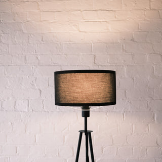 Close up of the Vanta floor lamp’s black KD fabric drum shade glowing softly against a white brick wall, showing its warm texture and modern finish.