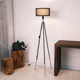 he Vanta floor lamp with a black KD fabric drum shade, matte black metal frame and gold dipped legs, styled in a minimalist room alongside natural wood stools, a dark timber desk and a potted plant. The soft lighting casts a warm glow against the white brick wall.