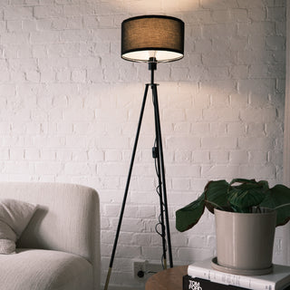 The Vanta floor lamp featuring a black KD fabric drum shade, matte black metal legs, and gold dipped ends. Shown in a modern living room beside a soft cream sofa and a wooden side table with a potted plant, styled against a white brick wall to highlight its warm ambient glow and contemporary design.