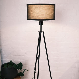 Close up of the Vanta floor lamp featuring a black KD fabric drum shade and matte black metal frame, styled against a white brick wall with a potted plant nearby.