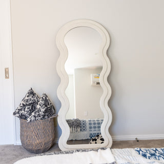 A stylish Velora Wavy Full-Length Mirror with a soft white velvet-textured frame, featuring elegant wavy edges. The mirror is leaned against a neutral-toned wall, reflecting a cosy bedroom setup with patterned cushions in a woven basket. Its contemporary design makes it a statement piece for modern interiors.