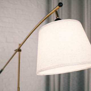 Close up view of the Vienna Brass Marble Base Floor Lamp showing the warm fabric lampshade and curved brass arm. The soft background of white brick and sheer curtains creates a calm, modern interior setting.
