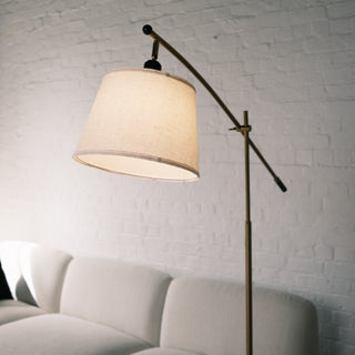 Close up of the Vienna Brass Marble Base Floor Lamp showing the warm fabric lampshade and elegant brass arm. The background features a soft white brick wall and a light textured sofa, creating a calm and modern interior look.