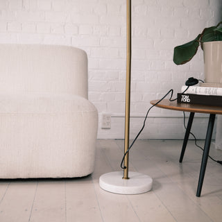 Close up of the Vienna Brass Marble Base Floor Lamp, showing the brushed brass pole and solid white marble base beside a cream textured sofa. A wooden side table with books and a plant sits in the background, styled in a modern New Zealand living room.
