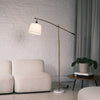 Vienna Brass Marble Base Floor Lamp standing beside a cream sofa with a white brick wall backdrop. The lamp features a brushed brass adjustable arm, off white fabric shade, and solid white marble base, styled in a modern New Zealand living room setting.