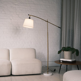 Vienna Brass Marble Base Floor Lamp standing beside a cream sofa with a white brick wall backdrop. The lamp features a brushed brass adjustable arm, off white fabric shade, and solid white marble base, styled in a modern New Zealand living room setting.