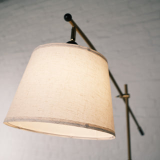 A close up view of the Vienna Brass Floor Lamp showing its warm textured linen shade and the curved brass arm behind it, photographed against a soft white brick wall for a clean modern look.
