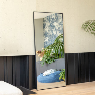 Zenith straight-edged full-length mirror (180 x 80 cm) with a slim 30 mm aluminium frame and ultra-clear 3 mm glass, leaning elegantly against a textured wall. The mirror reflects a bright, stylish living area with lush greenery and a blue bean bag, adding depth and sophistication to the contemporary interior.