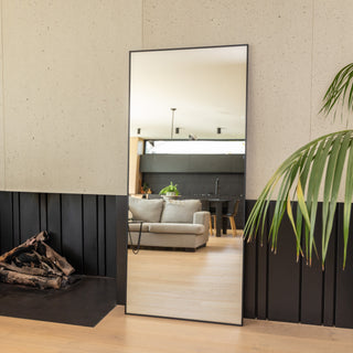 Zenith X full-length mirror (180 x 80 cm) with a sleek 30 mm black aluminium alloy frame and ultra-clear 3 mm glass, reflecting a bright modern living room with neutral tones, a grey sofa, and indoor plants. The mirror adds spaciousness and a refined aesthetic to the contemporary interior setting.