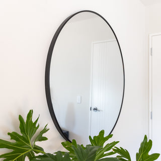 A sleek Zia Round Wall Mirror with a black aluminium alloy frame, mounted on a clean white wall. The mirror reflects a bright and airy interior, enhancing the room’s spacious and modern aesthetic. A lush green plant adds a natural contrast to the contemporary design. The thin metal frame and large circular shape make it a perfect statement piece for bedrooms, living rooms, or entryways. | C&F Creation